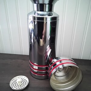 May include: A shiny, chrome cocktail shaker with red accents. The shaker is cylindrical with a flared top and a separate lid and strainer. The lid and base have red stripes. The strainer is silver with small holes.