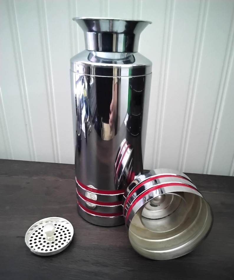 May include: A shiny, chrome cocktail shaker with red accents. The shaker is cylindrical with a flared top and a separate lid and strainer. The lid and base have red stripes. The strainer is silver with small holes.