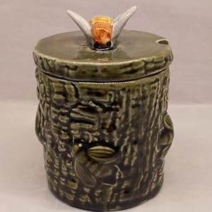 May include: A cylindrical ceramic jar with a dark green, textured tree bark finish. The lid features a sculpted bee with white wings and a yellow and orange body. A second bee is moulded into the side of the jar, which has a small notch for a spoon.