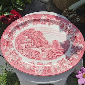 May include: An oval ceramic serving platter featuring a pink and white transferware pattern. The centre depicts a rural landscape with a house and trees, while the wide border is decorated with a detailed floral motif of roses and leaves.
