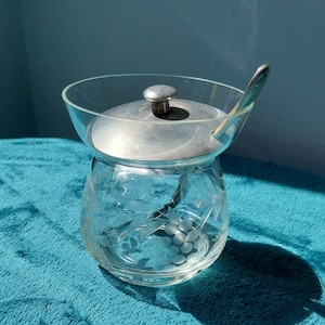 May include: A clear glass sugar bowl with an etched grape design and a silver lid. The bowl has a small spoon attached to the lid.