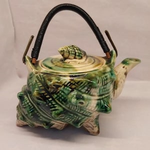 May include: A ceramic teapot shaped like a sea shell with a green and cream glaze. The lid features a small shell finial, and the handle is a black, coiled wire arch. The body has a textured, grid-like pattern with protruding spikes.