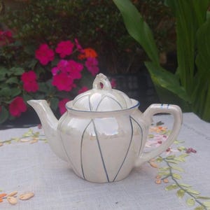 May include: A small, pearlescent white teapot with a lid, spout, and handle. The teapot has blue accent lines and a blue trim. The teapot is sitting on a white tablecloth with floral embroidery. The background features pink flowers and green foliage.