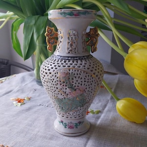 May include: White ceramic vase with a delicate floral design and intricate cutouts. The vase has gold accents and two small handles. It is sitting on a table with a floral pattern.