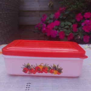 May include: A rectangular, pink plastic food container with a red lid. The container has a colourful illustration of tomatoes, peppers, and aubergines. It sits on a white tablecloth.