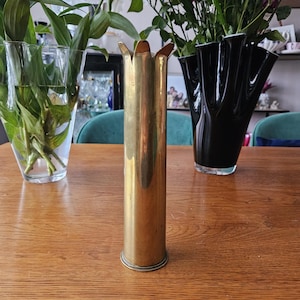 May include: A brass vase with a fluted top, on a wooden table. The vase is cylindrical with a petal-shaped opening. Other vases and flowers are in the background. The vase is approximately 25 cm tall.