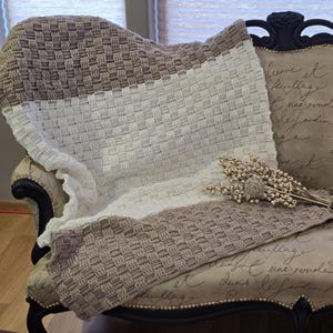 May include: A handmade crochet blanket in shades of white, beige, and brown, draped over a vintage armchair. The blanket features a woven basket weave pattern. A decorative floral arrangement rests on the chair.