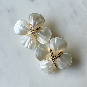 Peut inclure: Une paire de boucles d'oreilles en forme de fleur blanche avec des accents de fil d'or. Les boucles d'oreilles sont en nacre et ont un design délicat et féminin.