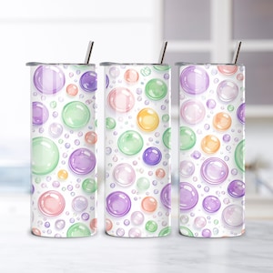 Puede incluir: Tres vasos de acero inoxidable con un diseño de burbujas de colores. Las burbujas son en tonos rosa, morado, verde, naranja y amarillo, sobre un fondo blanco. Cada vaso tiene una pajita plateada y una tapa.