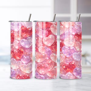 May include: Three stainless steel tumblers with a heart-themed design. The tumblers feature a pattern of red, pink, and purple hearts. Each tumbler has a silver straw and a lid. The background is a blurred kitchen setting.