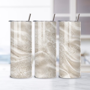 Op de afbeelding: Drie roestvrijstalen bekers met deksels en rietjes. De bekers hebben een beige, vloeiend stoffen ontwerp met delicate witte bloemenkantpatronen. Het ontwerp wikkelt zich om de cilindrische vorm van de bekers.
