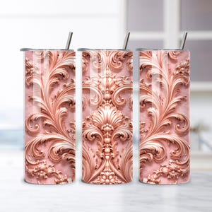 May include: Three stainless steel tumblers with a decorative rose gold baroque design. Each tumbler has a matching lid and a silver straw. The background is a blurred image of a kitchen.