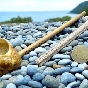 May include: A handcrafted smoking pipe with a seashell bowl, a wooden stem with "CHALWAH" etched on it, a textured filter, and two round screens. The pipe is displayed on a bed of smooth, gray and blue stones.