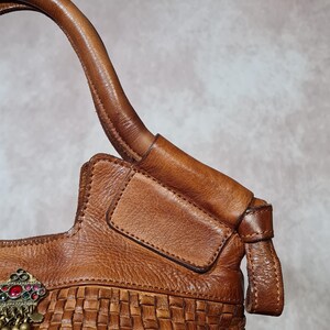Peut inclure: Sac &agrave; main en cuir marron avec une base tress&eacute;e et un fermoir d&eacute;coratif en m&eacute;tal avec des accents rouges, verts et dor&eacute;s. Le sac a une poign&eacute;e incurv&eacute;e et un rabat. Le cuir semble vieilli.