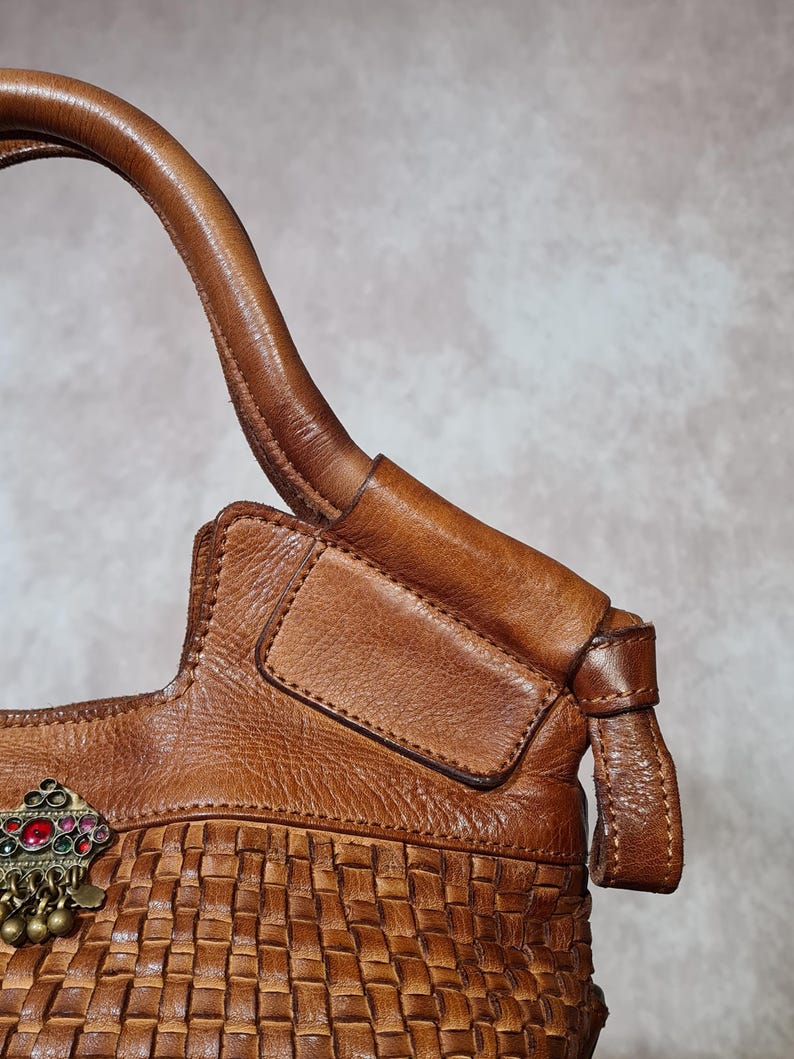 Peut inclure: Sac &agrave; main en cuir marron avec une base tress&eacute;e et un fermoir d&eacute;coratif en m&eacute;tal avec des accents rouges, verts et dor&eacute;s. Le sac a une poign&eacute;e incurv&eacute;e et un rabat. Le cuir semble vieilli.