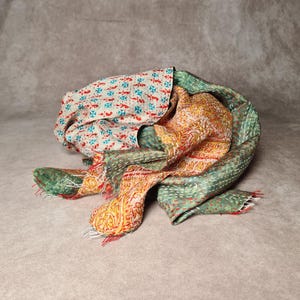 May include: A colorful, patterned scarf with a mix of floral and geometric designs. The scarf features a cream-colored fabric with red and blue motifs, alongside sections of green and orange with intricate patterns. The edges are finished with red fringe.