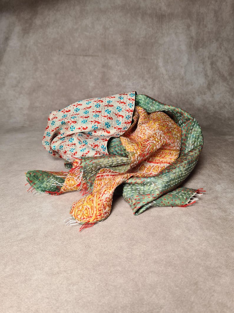 May include: A colorful, patterned scarf with a mix of floral and geometric designs. The scarf features a cream-colored fabric with red and blue motifs, alongside sections of green and orange with intricate patterns. The edges are finished with red fringe.