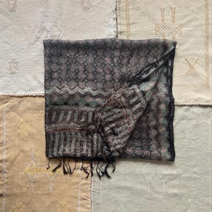 May include: A dark patterned scarf with a black border and fringe. The scarf features a repeating floral design in shades of black, brown, and teal. The scarf is folded and draped, showcasing its intricate pattern and texture.