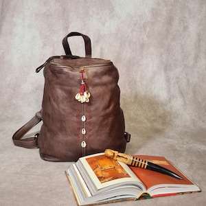 May include: A brown leather backpack with a top handle, zipper closure, and adjustable straps. The backpack features decorative stitching and a tassel with shells. An open book and a decorative pen rest on a surface.