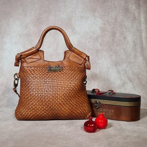 Peut inclure: Un sac &agrave; main en cuir marron avec un motif tiss&eacute;, dot&eacute; d'une poign&eacute;e sup&eacute;rieure et d'un fermoir d&eacute;coratif. &Agrave; c&ocirc;t&eacute;, deux petits ornements en verre rouge en forme de grenade et une bo&icirc;te en bois avec un couvercle d&eacute;coratif.