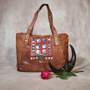 Könnte beinhalten: Eine braune Ledertasche mit einem zentralen rechteckigen Einsatz, der mit runden Spiegeln, bunter Stoffstickerei und hängenden Kaurimuscheln verziert ist. Eine dunkle Mondsichelskulptur und eine einzelne rosa Rose liegen davor.