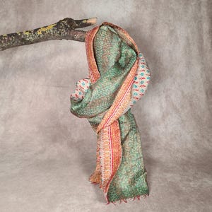 May include: A green and red patterned scarf draped over a tree branch. The scarf has a detailed design with a mix of colors, including red, green, and white. The edges are finished with a fringe.