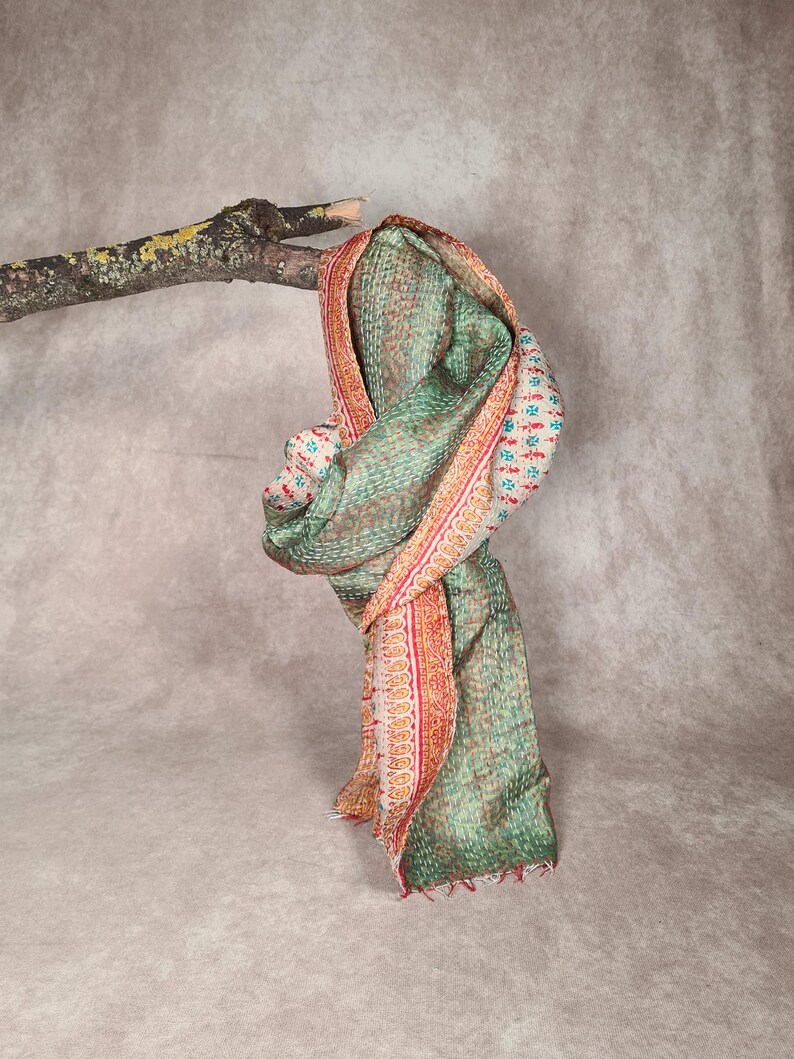 May include: A green and red patterned scarf draped over a tree branch. The scarf has a detailed design with a mix of colors, including red, green, and white. The edges are finished with a fringe.