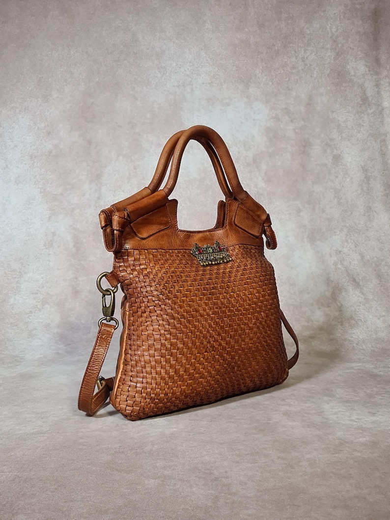 Peut inclure: Un sac &agrave; main en cuir marron avec un motif tiss&eacute;. Le sac poss&egrave;de deux poign&eacute;es et une bandouli&egrave;re amovible. Un embl&egrave;me d&eacute;coratif en m&eacute;tal est pr&eacute;sent sur le devant. Le sac est en cuir et a un look classique et intemporel.