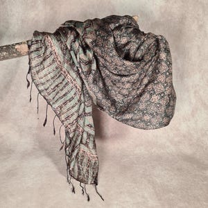 May include: A patterned scarf in shades of green, pink, and black is draped over a wooden branch. The scarf features a mix of floral and striped designs, with fringed edges. The fabric appears soft and lightweight, suitable for various styles.