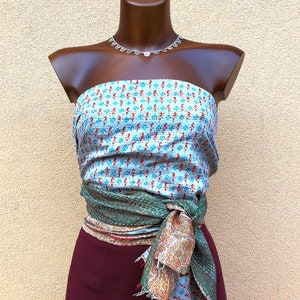May include: A mannequin displays a layered outfit. A light blue and red patterned top is paired with a green and patterned wrap tied at the waist. A maroon skirt completes the look. A silver necklace is also included.
