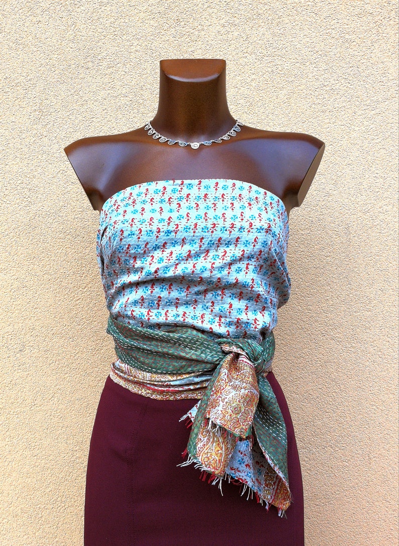 May include: A mannequin displays a layered outfit. A light blue and red patterned top is paired with a green and patterned wrap tied at the waist. A maroon skirt completes the look. A silver necklace is also included.