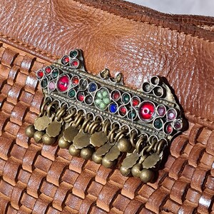 Peut inclure: Une broche orn&eacute;e en m&eacute;tal argent&eacute;, orn&eacute;e de pierres pr&eacute;cieuses color&eacute;es et de breloques en laiton pendantes. La broche pr&eacute;sente des pierres rouges, bleues, vertes et transparentes. Le fond est une surface en cuir tress&eacute; marron.