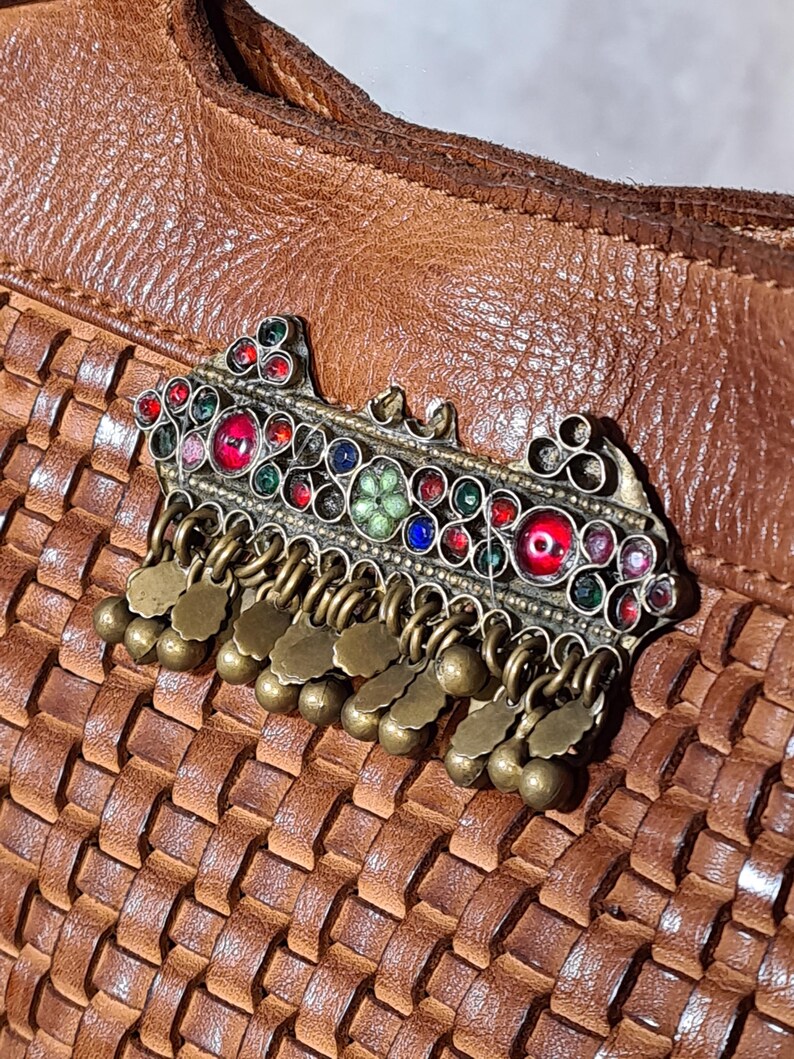 Peut inclure: Une broche orn&eacute;e en m&eacute;tal argent&eacute;, orn&eacute;e de pierres pr&eacute;cieuses color&eacute;es et de breloques en laiton pendantes. La broche pr&eacute;sente des pierres rouges, bleues, vertes et transparentes. Le fond est une surface en cuir tress&eacute; marron.