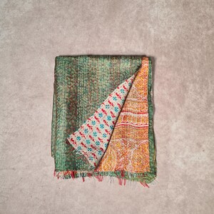 May include: A folded, reversible textile with a green, stitched exterior and a patterned interior. The interior features a white background with blue and red floral designs on one side and a yellow and orange paisley pattern on the other. The edges are fringed.