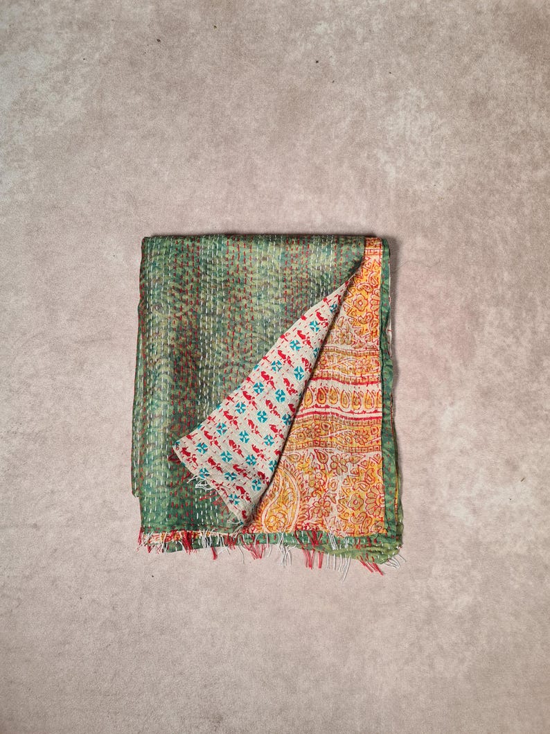 May include: A folded, reversible textile with a green, stitched exterior and a patterned interior. The interior features a white background with blue and red floral designs on one side and a yellow and orange paisley pattern on the other. The edges are fringed.