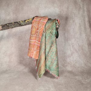 May include: Two colorful, patterned scarves draped over a tree branch. One scarf features a warm orange and pink paisley design, while the other is a green and pink stitched pattern. The scarves have fringed edges.