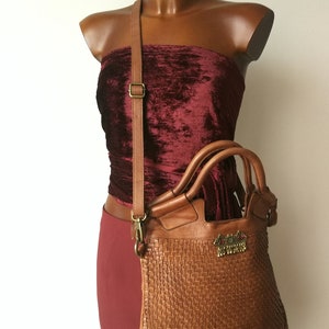 Peut inclure: Sac &agrave; main en cuir tress&eacute; marron avec bandouli&egrave;re et poign&eacute;es sup&eacute;rieures. Le sac comporte un embl&egrave;me d&eacute;coratif et est pr&eacute;sent&eacute; sur un mannequin portant un haut et une jupe en velours bordeaux. Dimensions du sac non visibles.