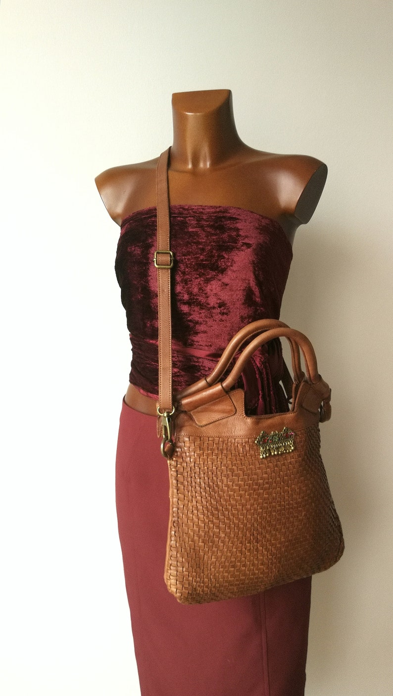 Peut inclure: Sac &agrave; main en cuir tress&eacute; marron avec bandouli&egrave;re et poign&eacute;es sup&eacute;rieures. Le sac comporte un embl&egrave;me d&eacute;coratif et est pr&eacute;sent&eacute; sur un mannequin portant un haut et une jupe en velours bordeaux. Dimensions du sac non visibles.