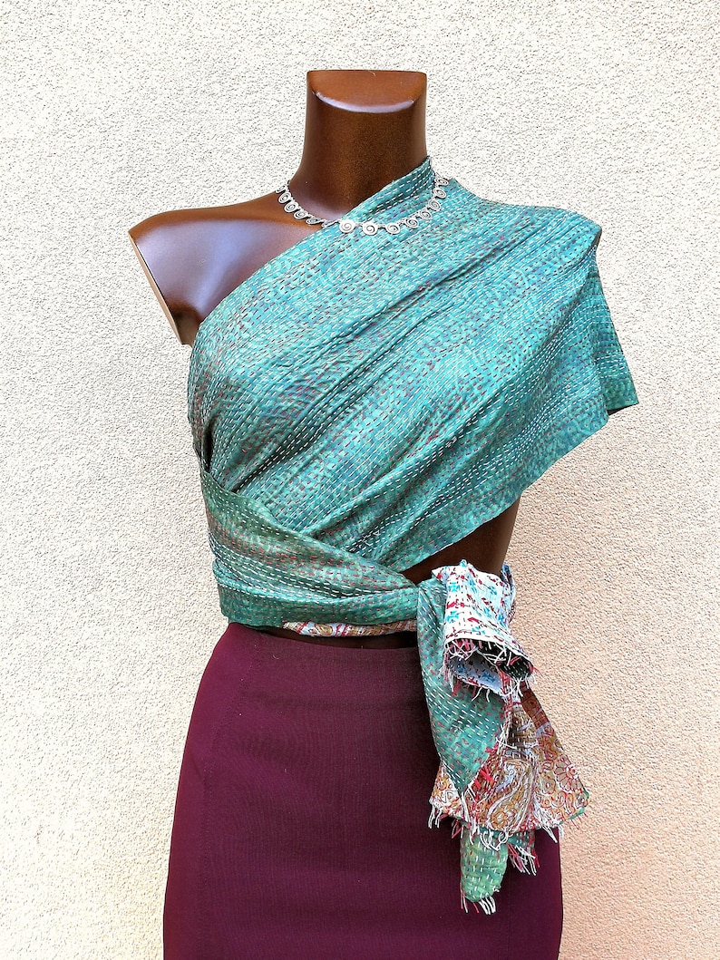 May include: A teal and brown patterned wrap is draped over a mannequin, paired with a burgundy skirt. The wrap is secured at the waist, with a fringed edge revealing a colorful paisley design. A silver necklace adorns the mannequin's neck.