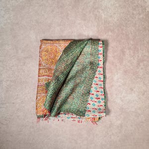 May include: Folded textile with a patchwork design. The top layer is a green fabric with a quilted pattern. Beneath it, a section of yellow fabric with a floral pattern is visible, along with a section of white fabric with red and blue floral patterns.