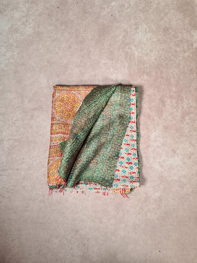 May include: Folded textile with a patchwork design. The top layer is a green fabric with a quilted pattern. Beneath it, a section of yellow fabric with a floral pattern is visible, along with a section of white fabric with red and blue floral patterns.