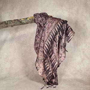 May include: A patterned scarf in shades of purple and black is draped over a tree branch. The scarf features an intricate design with floral and geometric elements. The fabric appears lightweight and flowing, with a delicate texture. The scarf's edges are finished with small tassels.