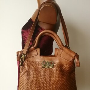 Peut inclure: Sac fourre-tout en cuir marron avec un motif tiss&eacute;. Le sac est orn&eacute; d'une d&eacute;coration m&eacute;tallique avec des accents rouges et verts. Il est dot&eacute; d'une bandouli&egrave;re et de deux poign&eacute;es sup&eacute;rieures.