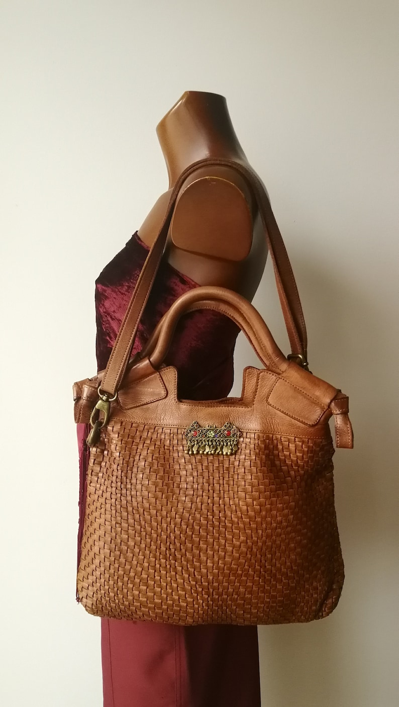 Peut inclure: Sac fourre-tout en cuir marron avec un motif tiss&eacute;. Le sac est orn&eacute; d'une d&eacute;coration m&eacute;tallique avec des accents rouges et verts. Il est dot&eacute; d'une bandouli&egrave;re et de deux poign&eacute;es sup&eacute;rieures.