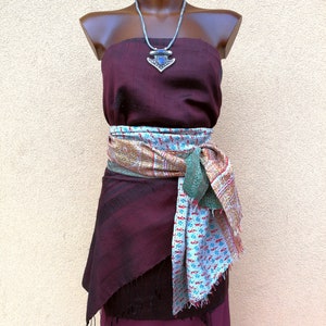 May include: A burgundy strapless dress with a patterned scarf tied around the waist. A silver necklace with a blue pendant is displayed on a mannequin. The dress has a layered skirt with fringe.