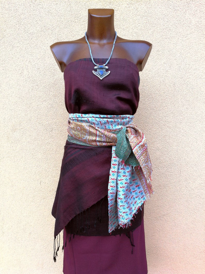 May include: A burgundy strapless dress with a patterned scarf tied around the waist. A silver necklace with a blue pendant is displayed on a mannequin. The dress has a layered skirt with fringe.