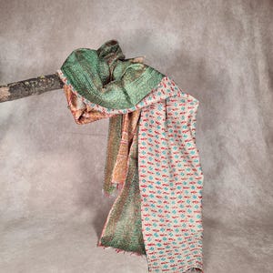 May include: A multi-colored scarf draped over a wooden branch. The scarf features a green patterned section, a section with orange and red patterns, and a section with a white background and red and teal floral designs. The scarf has a fringed edge.