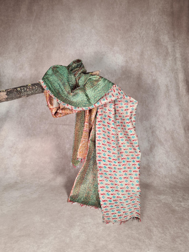 May include: A multi-colored scarf draped over a wooden branch. The scarf features a green patterned section, a section with orange and red patterns, and a section with a white background and red and teal floral designs. The scarf has a fringed edge.