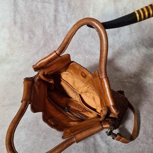 Peut inclure: Sac &agrave; main en cuir marron avec une poign&eacute;e incurv&eacute;e. Le sac pr&eacute;sente un panneau lat&eacute;ral tiss&eacute; et une poche int&eacute;rieure zipp&eacute;e. L'int&eacute;rieur du sac est d'une teinte marron plus claire.