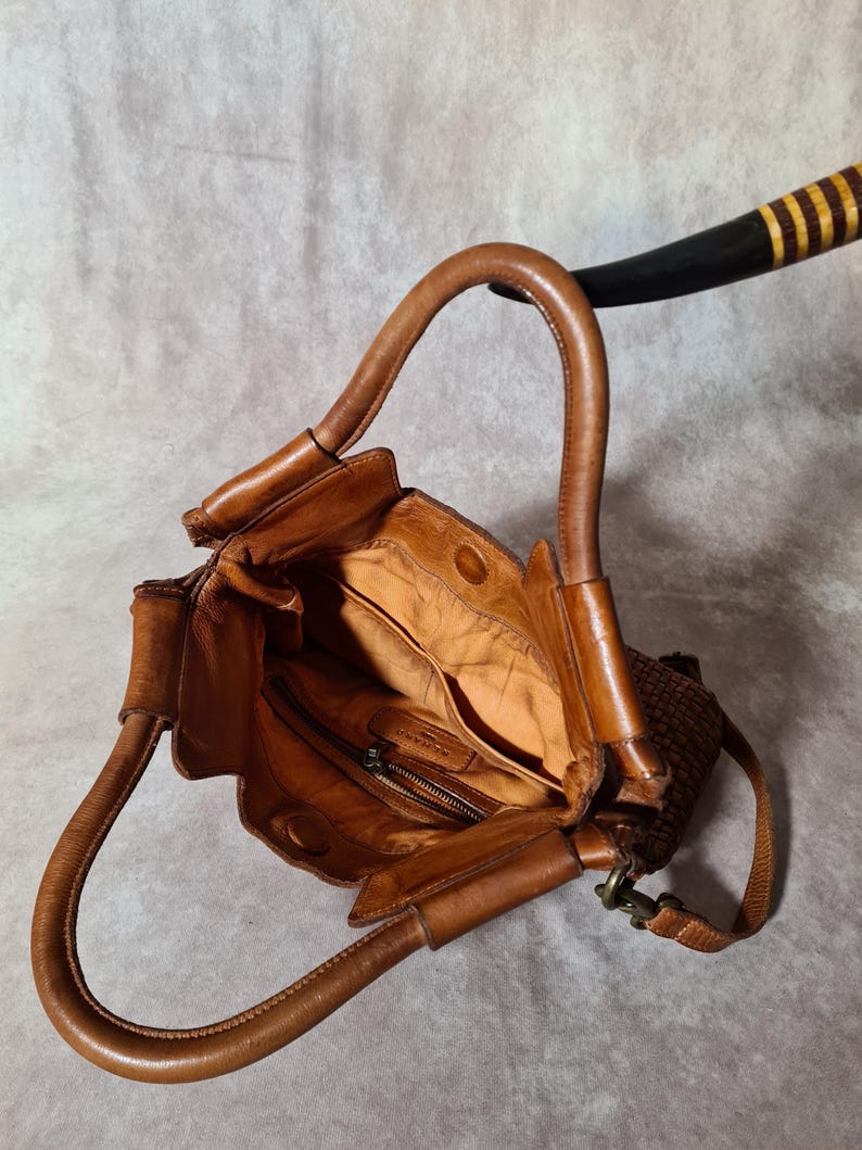 Peut inclure: Sac &agrave; main en cuir marron avec une poign&eacute;e incurv&eacute;e. Le sac pr&eacute;sente un panneau lat&eacute;ral tiss&eacute; et une poche int&eacute;rieure zipp&eacute;e. L'int&eacute;rieur du sac est d'une teinte marron plus claire.